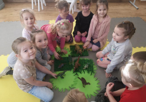 Widok na grupę dziewczynek, które układają prehistoryczny las wykorzystując plastikowe dinozaury, zielone serwetki, trawki.